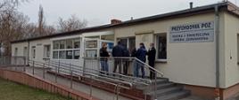 Zdjęcie przedstawia budynek Przychodni POZ, gdzie odbywają się szczepienia przeciwko COVID-19. Na schodach przed wejściem do budynku stoi grupa mężczyzn. Przed budynkiem widać trawnik, a za nim po lewej stronie rosnące drzewa.