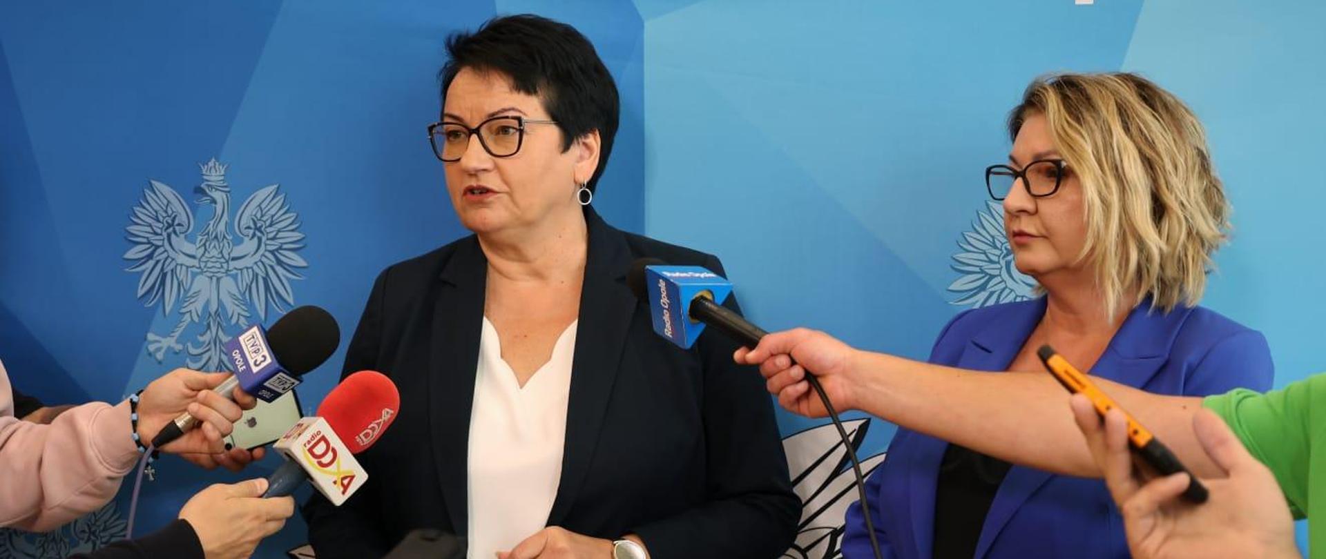 Wojewoda Opolski Monika Jurek oraz Dyrektor Wydziału Polityki Społecznej Ewa Pawlinów w trakcie konferencji prasowej dotyczącej programu "Opieka 75+". Na zdjęciu widać, jak Wojewoda mówi do mikrofonów trzymanych przez dziennikarzy. Obok, po prawej stronie stoi Dyrektor Ewa Pawlinów. 