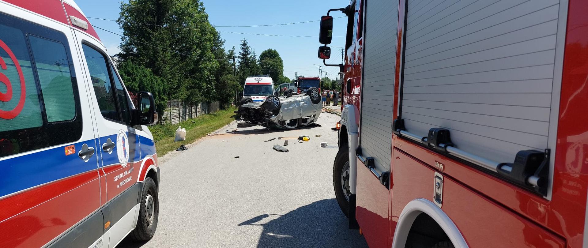 Zdjęcie na pierwszym planie przedstawia ustawione obok siebie dwa pojazdy- samochód straży pożarnej oraz karetkę pogotowia. W tle widać uszkodzony pojazd osobowy znajdujący się na dachu i całkowicie blokujący ruch w obu kierunkach drogi powiatowej. Na drodze porozrzucane elementy uszkodzonego samochodu. Za nim widać kolejną karetkę pogotowia oraz jeszcze jeden samochód ratowniczo-gaśniczy.