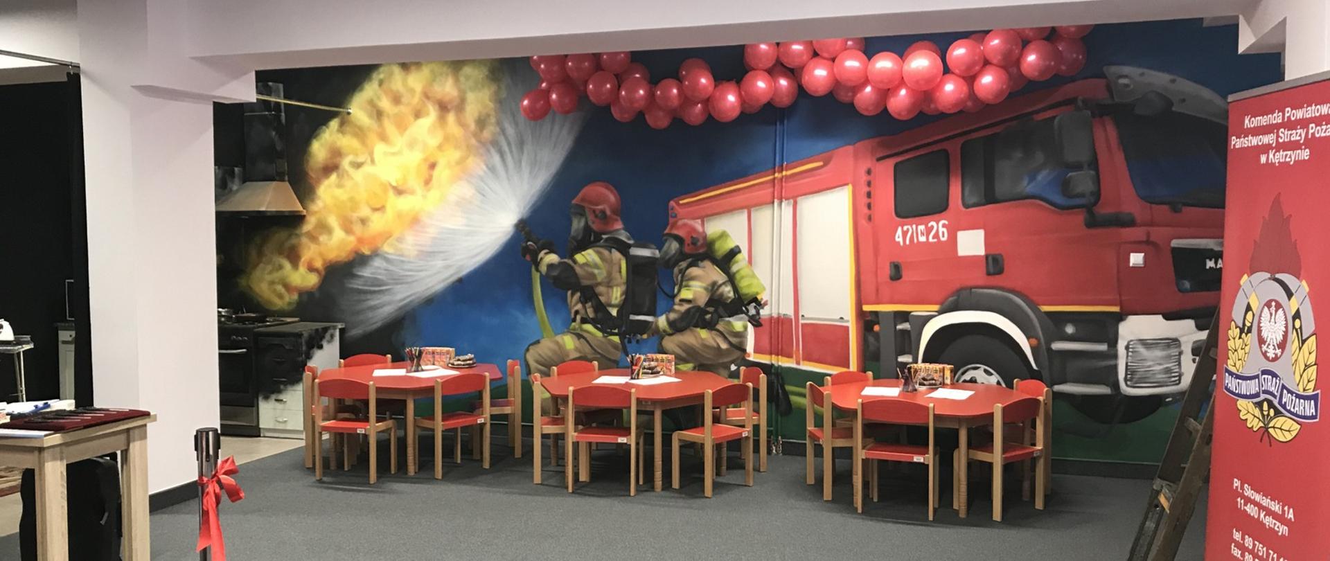 Zdjęcie zrobione w Sali Edukacyjnej w Kętrzynie. Stoliki z krzesełkami dla dzieci oraz ściana z namalowanym muralem przedstawiającym wóz strażacki, dwóch strażaków ubranych w pełne ubranie bojowe oraz ogień wydobywający się z kuchni budynku.
