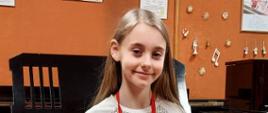 Zdjęcie kolorowe. Dziewczynka - Alicja Puchalik w ręku trzyma dyplom konkursu i pluszową zabawkę w formie poduszki. W tle fortepian .