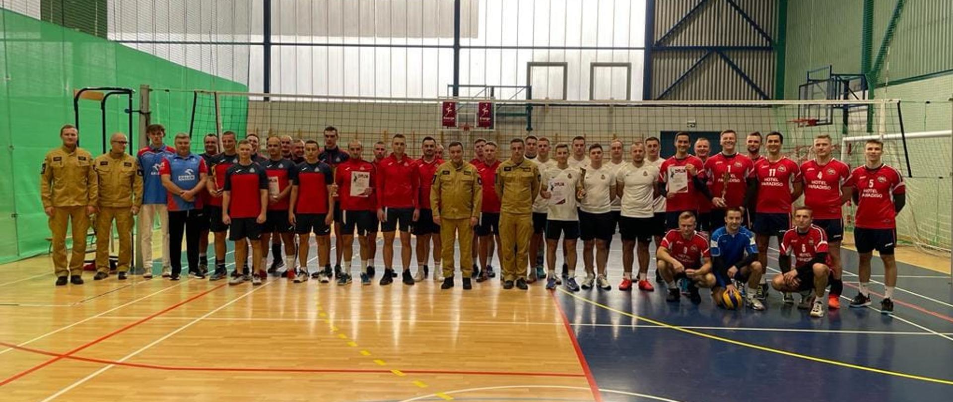 Na zdjęciu widzimy wszystkich uczestników oraz organizatorów zawodów sportowych rozgrywanych w hali sportowej.