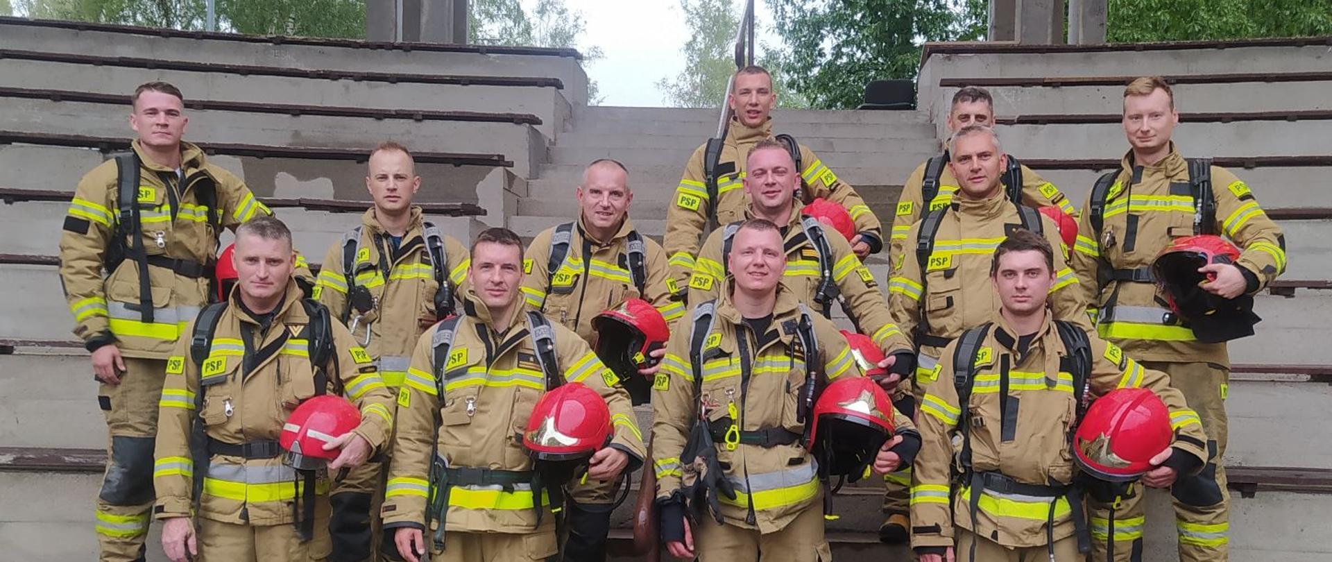 Na tle schodów stoi kilku strażaków w piaskowych mundurach bojowych w rękach trzymają czerwone hełmy.