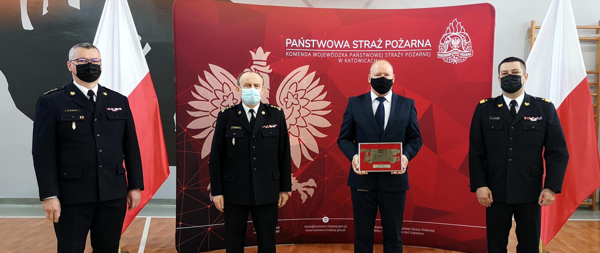 Na zdjęciu widoczna jest sala w Komendzie Wojewódzkiej PSP w Katowicach. Na tle dwóch flag państwowych i ścianki z godłem Polski i napisem Państwowa Straż Pożarna Komenda Wojewódzka PSP w Katowicach, od prawej stoi Śląski Komendant Wojewódzki PSP w mundurze wyjściowym i maseczce ochronnej, Pan Andrzej Heród w garniturze i maseczce ochronnej, oraz zastępcy śląskiego Komendanta wojewódzkiego st. bryg. Bogdan Jędrocha i st. bryg. Mirosław Synowiec, obydwaj w mundurach wyjściowych i maseczkach ochronnych. 