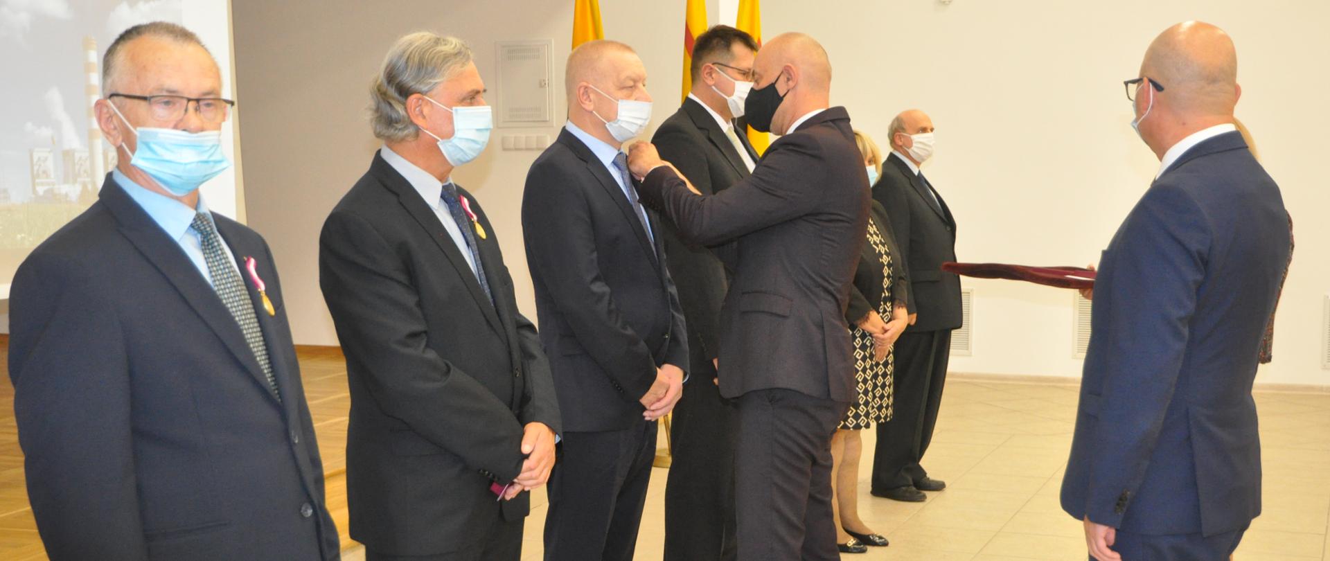 Wojewoda Opolski wręcza zasłużonym pracownikom Elektrowni Opole odznaczenia państwowe. W kadrze sześcioro osób stoi w rzędzie i oczekuje na wręczenie odznaczenia. Po prawej stronie widać sylwetkę mężczyzny skierowanego bokiem oraz kobietę trzymająca tace z odznaczeniami. 