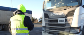 Osoba ubrana w odblaskową kamizelkę z napisem Inspekcja Ochrony Środowiska stojąca tyłem do zdjęcia. Obok osoby stoi zaparkowany samochód ciężarowy z napisem odpady.