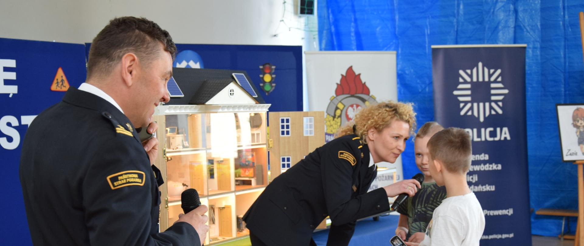 Strażak daje mikrofon chłopcom do mówienia, obok stoi drugi funkcjonariusz. Za nimi stoi mobilny symulator zagrożeń pożarowych.