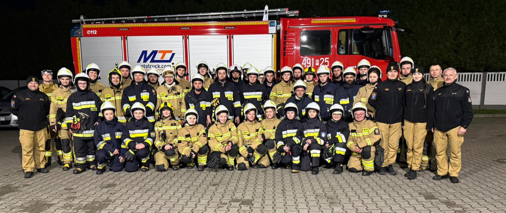 Zdjęcie przedstawia druhów OSP oraz funkcjonariuszy PSP na tle pojazdu pożarniczego po zmroku