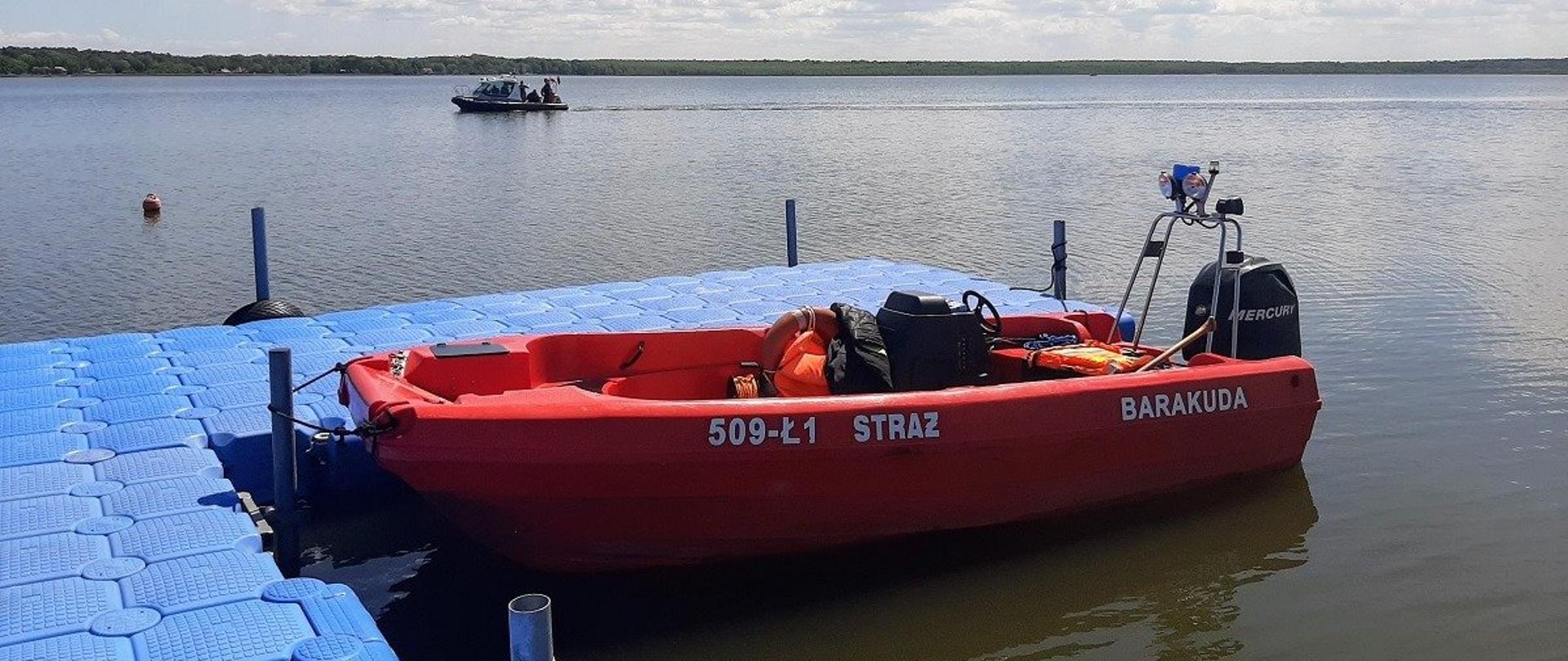 Na zdjęciu przycumowana do niebieskiego pływającego pomostu pomarańczowa łódź motorowa z kadłubem wykonanym z tworzywa sztucznego. Widoczna lewa burta z białymi napisami: 509 Ł1, Straż, Barakuda. Na burcie silnik motorowy. W tle otwarty akwen z przepływającą łodzią, dalej na horyzoncie las. Niebo pogodne błękitne z niewielkimi obłokami.