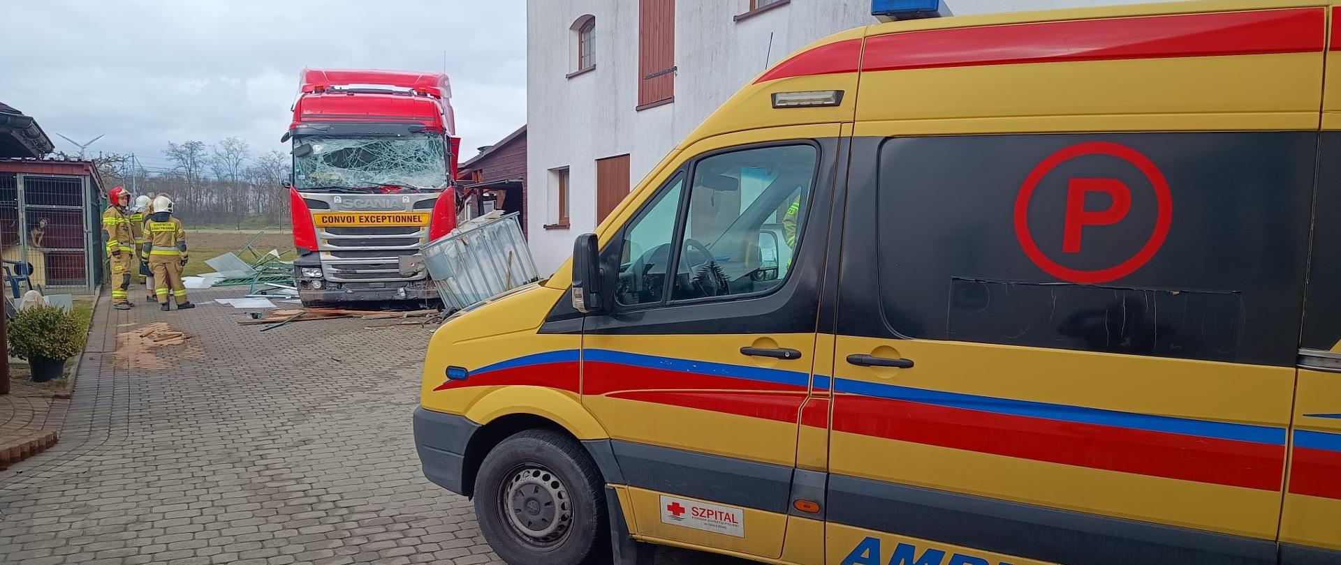 Na zdjęciu widać ciężarówkę z rozbitą przednią szybą, która zatrzymała się na prywatnej posesji tuż przy budynku, niszcząc ogrodzenie i elementy zabudowy. Obok stoją strażacy w ubraniach ochronnych, oceniający sytuację i zabezpieczający miejsce zdarzenia. Na pierwszym planie zaparkowany jest ambulans.