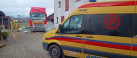 Na zdjęciu widać ciężarówkę z rozbitą przednią szybą, która zatrzymała się na prywatnej posesji tuż przy budynku, niszcząc ogrodzenie i elementy zabudowy. Obok stoją strażacy w ubraniach ochronnych, oceniający sytuację i zabezpieczający miejsce zdarzenia. Na pierwszym planie zaparkowany jest ambulans.
