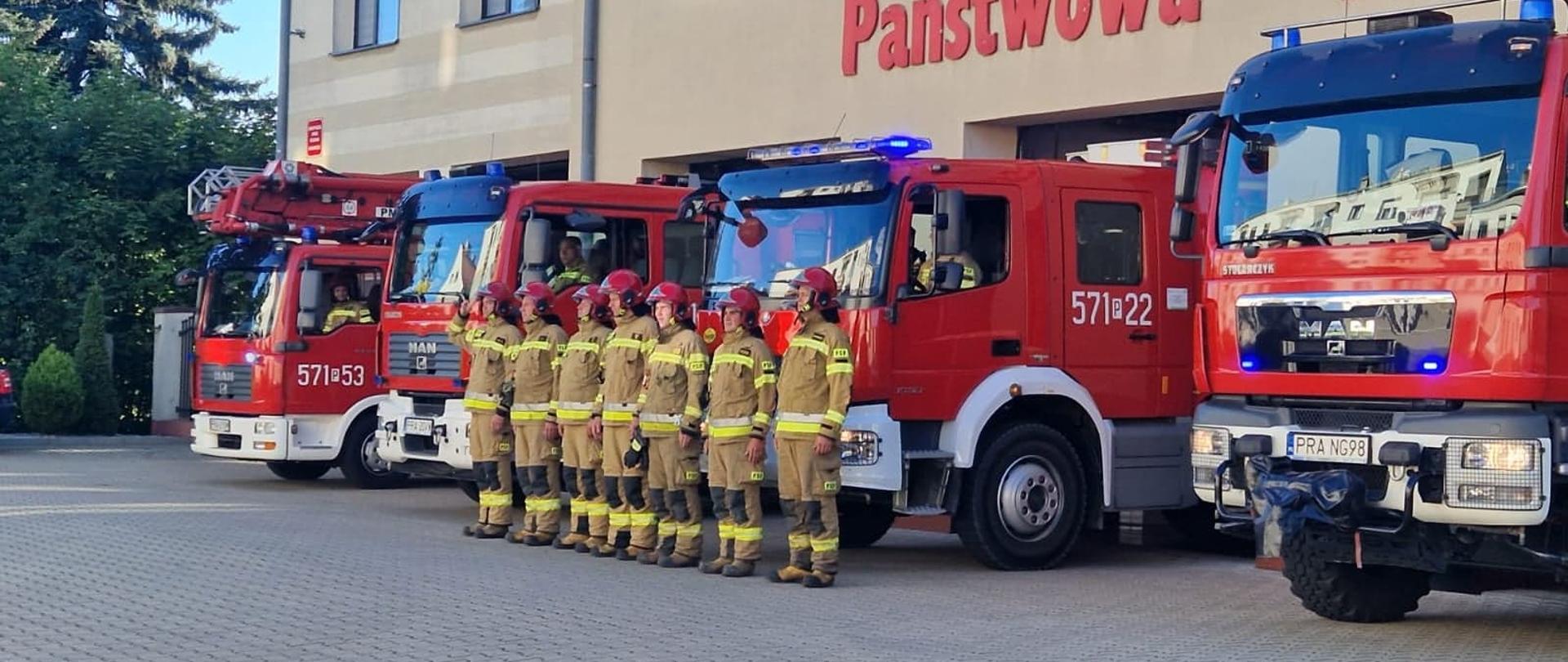 Uczczenie pamięci tragicznie zmarłego strażaka. Plac przed Komendą Powiatową PSP w Rawiczu. Przed garażami stoją 4 pojazdy strażackie z włączonymi sygnałami świetlnymi. Przed nimi 7 strażaków JRG ustawionych w szeregu, w ubraniach specjalnych i czerwonych hełmach. Dowódca salutuje.