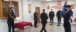 Na zdjęciu w mundurach galowych po lewej stronie Zastępca Komendanta Powiatowego a po prawej poczet sztandarowy ze sztandarem. Strażacy składający ślubowanie na środku w ubraniach czarnych koszarowych. Komendant Powiatowy podchodzi do nich i wręcza akty ślubowania.Uroczystość ślubowania mam miejsce w sali konferencyjnej.