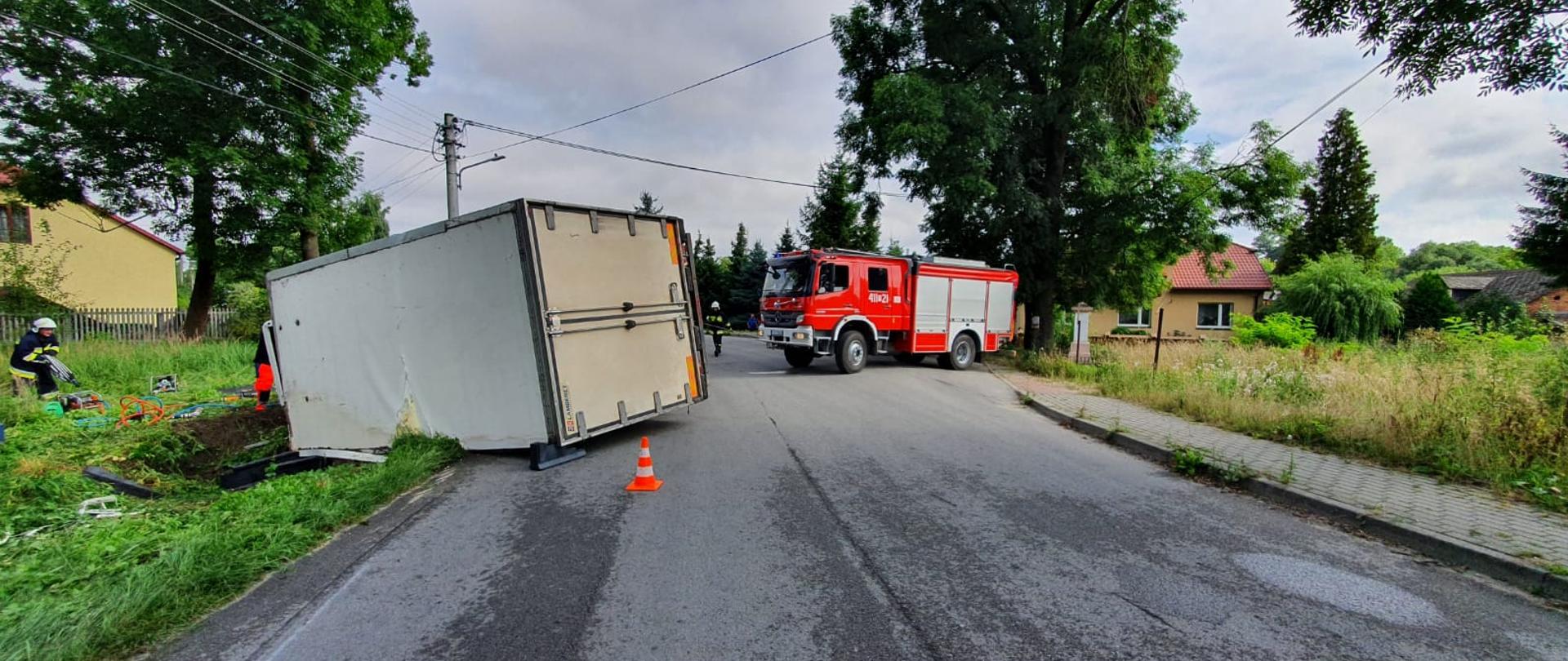 Zdjęcie przedstawia przewrócony samochód ciężarowy na lewy bok, częściowo znajdujący się na jezdni, częściowo w rowie. Na drugim planie widnieje samochód ratowniczo-gaśniczy. Po lewej stronie strażak w umundurowaniu specjalnym rozwija sprzęt hydrauliczny. 