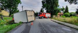 Zdjęcie przedstawia przewrócony samochód ciężarowy na lewy bok, częściowo znajdujący się na jezdni, częściowo w rowie. Na drugim planie widnieje samochód ratowniczo-gaśniczy. Po lewej stronie strażak w umundurowaniu specjalnym rozwija sprzęt hydrauliczny. 