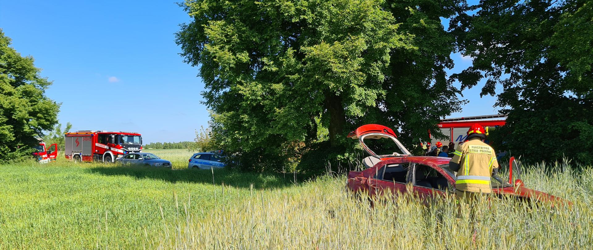 Zdjęcie przedstawia pole i znajdujący się w nim czerwony samochód z otwartym bagażnikiem i drzwiami, trochę porysowany i z wgnieceniami, przy samochodzie stoi strażak w piaskowym stroju strażackim. Z tyłu widać drzewa, a przy nich drogę, na której stoją 3 samochody straży pożarnej i 1 policji.