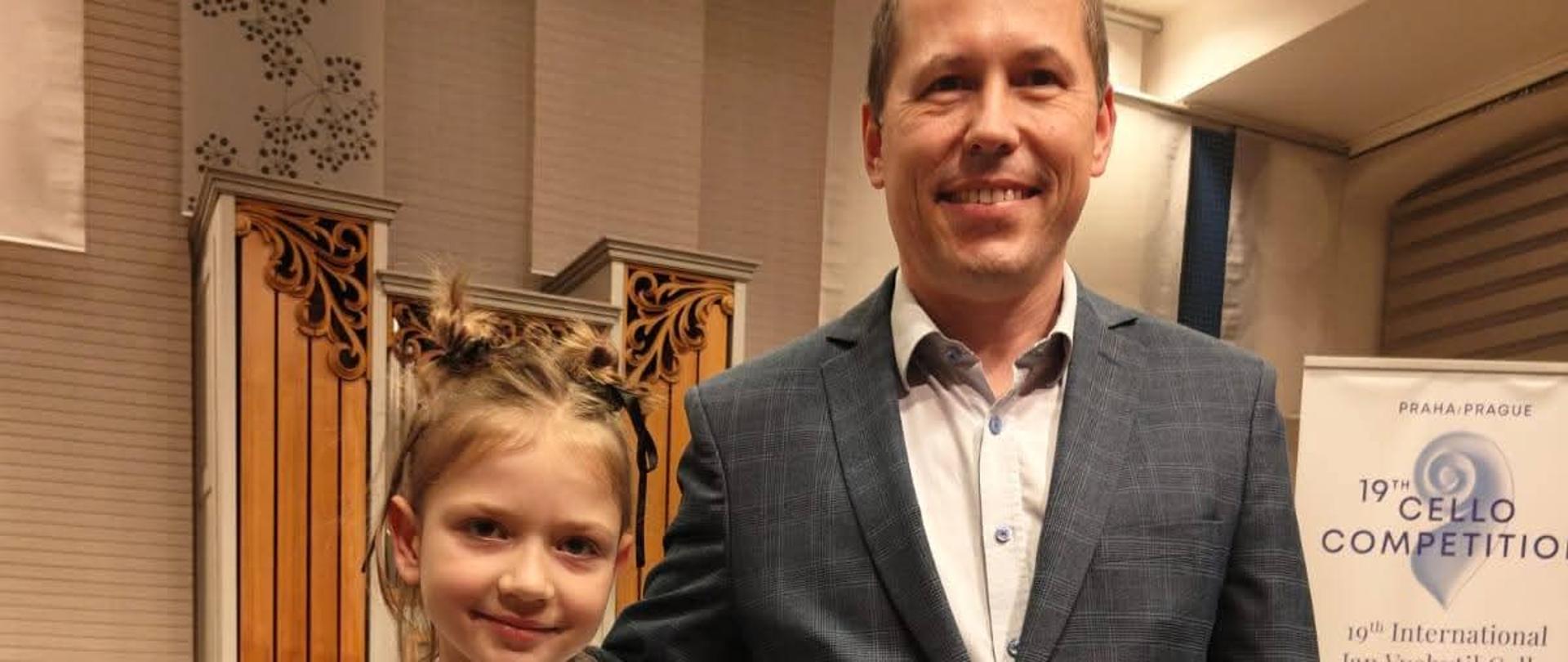 na zdjęciu po lewej stronie dziewczyna około dziesiecioletnia z w odświetnym ubraniu trzymająca dyplom a na szyi zawieszony ma medal na błękitnej wstążce, po prawej stronie stoi nauczyciel w czarnych spodniach i szarej marynarce