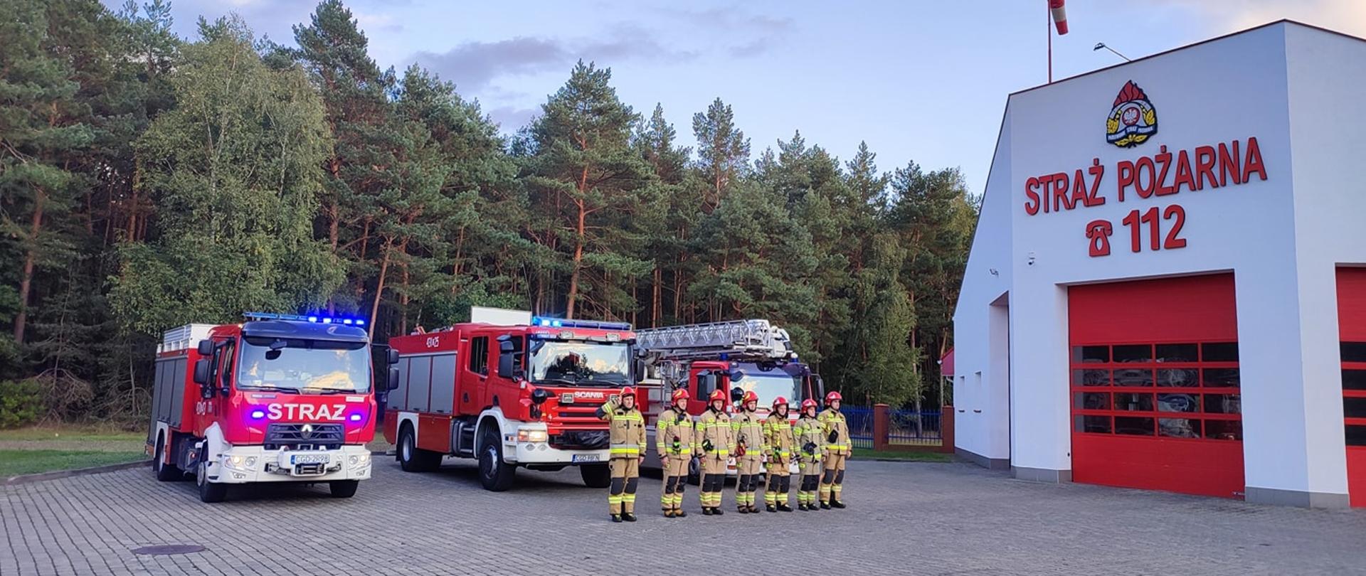 STRAŻACY W UBRANIACH SPECJALNYCH STOJĄ USTAWIENI W SZEREGU NA TLE POJAZDÓW RATOWNICZO-GAŚNICZYCH.