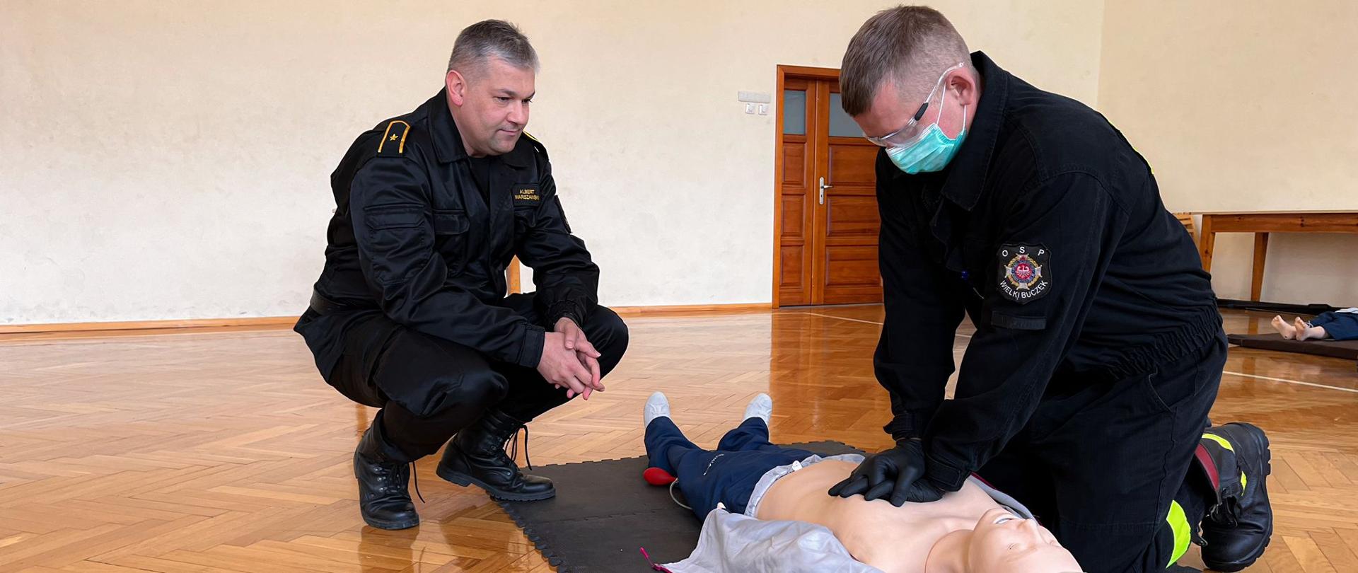 Strażacy w ubraniach koszarowych z maseczkami i okularami wykonują działania z zakresu kwalifikowanej pierwszej pomocy na manekinie. Zdjęcie wykonane na sali gimnastycznej budynku strażnicy.