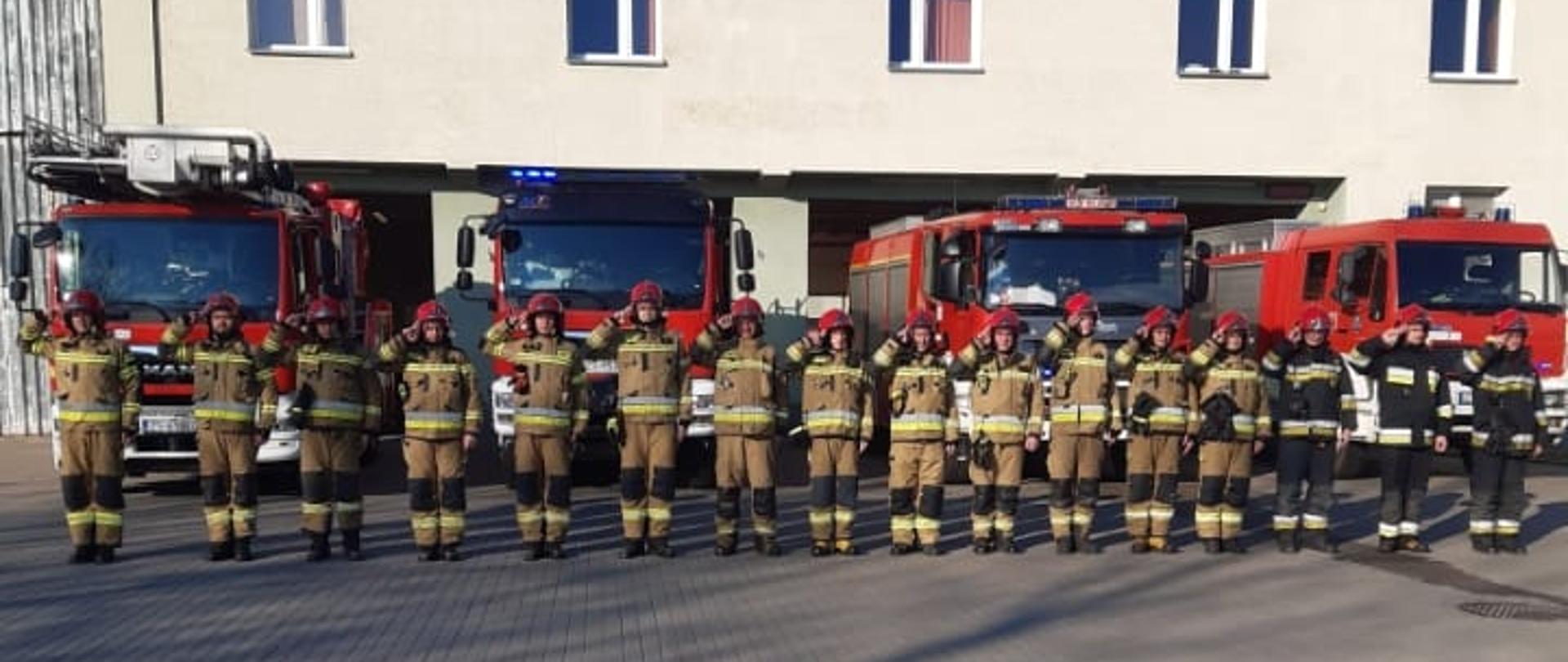 Minuta ciszy dla bohaterskich ratowników Ukrainy, strażacy ubrani w ubrania specjalne koloru piaskowego oraz granatowego stoją na tle samochodów specjalnych.