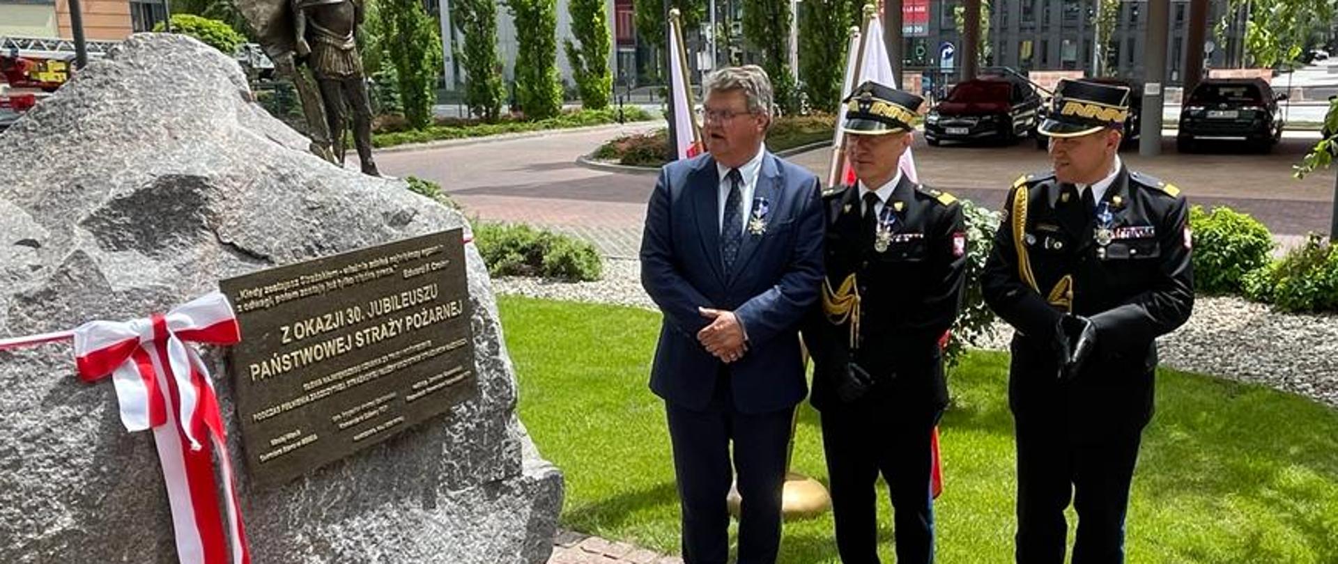 Wiceminister MSWiA oraz komendant główny PSP wraz z zastępcą w trakcie odsłonięcia pamiątkowego kamienia tablicą jubileuszową. 