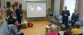 Sala wykładowa z seniorami w tle wyświetlany film na rzutniku, burmistrz i funkcjonariusze PSP
