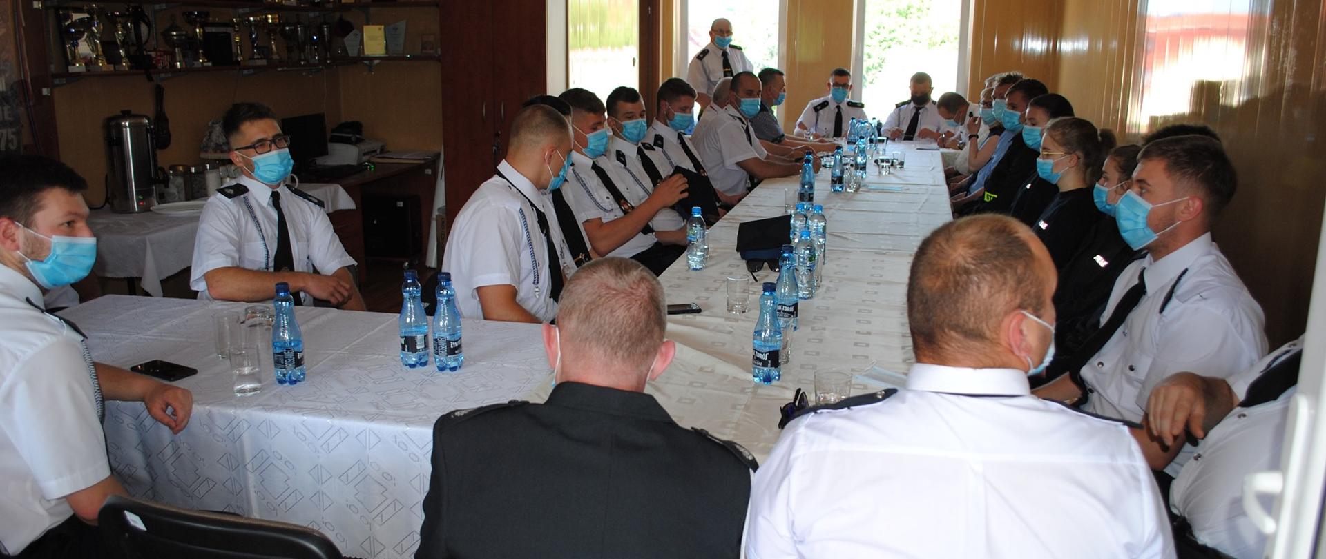 Na zdjęciu pomieszczenie, w którym przy stole okrytym białym obrusem ustawionym w literę L siedzą strażacy w białych koszulach z krótkim rękawkiem, nieliczni w ciemnych bluzach i maseczkach na twarzach. Na stole butelki z wodą. W tle półki z pucharami.