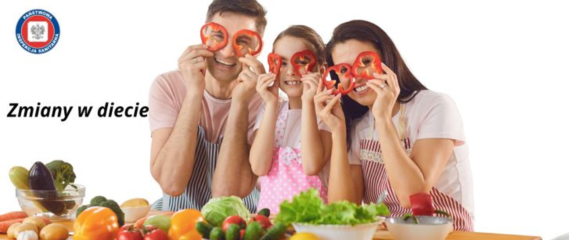 Kobieta, mężczyzna i dziewczynka trzymający przy twarzy plastry papryki stoją przy stole na którym położono warzywa