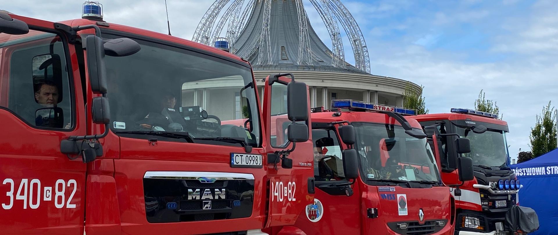 Na pierwszym planie trzy pojazdy pożarnicze, na tle kościoła. Ekspozycja w miasteczku strażackim.