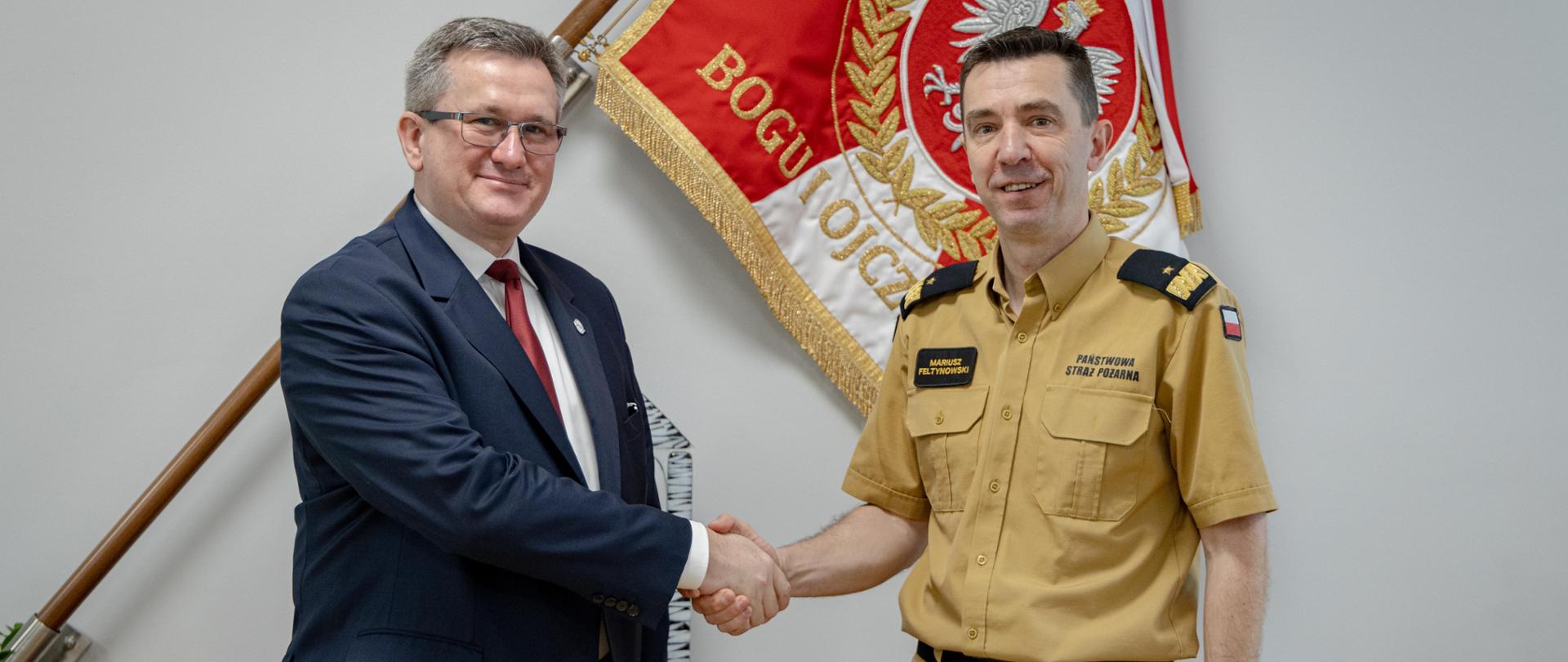 Komendant główny PSP ściska dłoń z mężczyzną w garniturze. W tle sztandar komendy głównej.