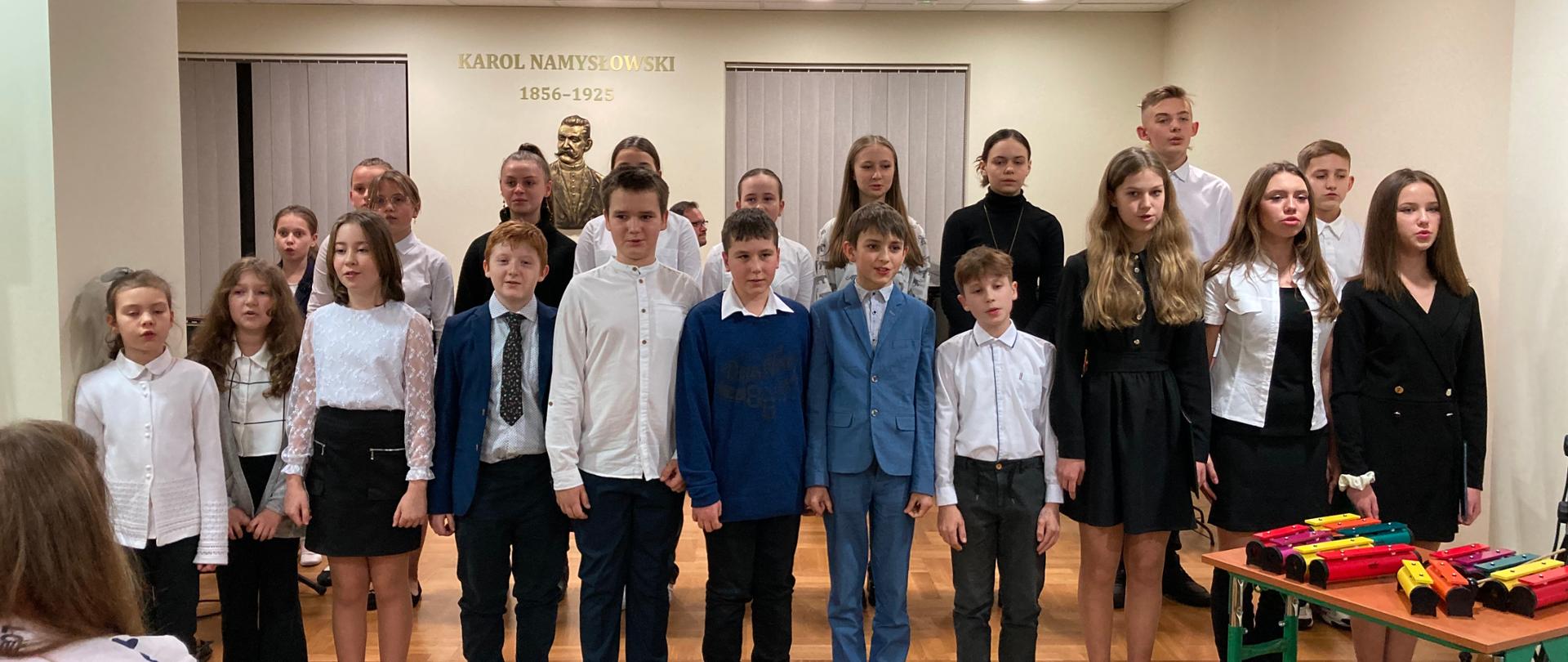 Zdjęcie - koncert kolęd. Scena sali koncertowej szkoły muzycznej. Z tyłu popiersie Karola Namysłowskiego, po prawej stronie choinka. Na pierwszym planie chór szkolny. 