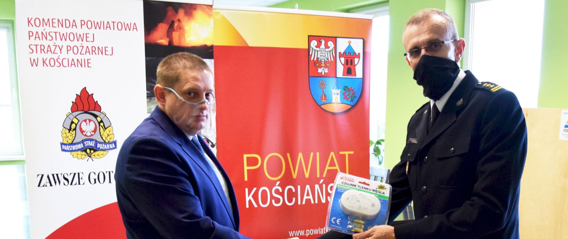 Kolejny rok powiat funduje czujki dla mieszkańców. Na zdjęciu wicestarosta powiatu kościańskiego przekazuje symbolicznie czujkę tlenku węgla na ręce Komendanta Powiatowego PSP w Kościanie. W tle czujki tlenku węgla i banery jednostek.