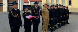 Na zdjęciu widoczna jest grupa osób w mundurach strażackich stojących w szeregu na placu, przed budynkiem straży pożarnej. Trzech mężczyzn z przodu ubranych jest w galowe mundury strażackie, jeden z nich trzyma złożoną polską flagę. Obok nich stoi osoba w beżowym mundurze, a za nimi ustawiona jest grupa strażaków w czarnych mundurach z żółtymi odblaskami. W tle widoczne są budynki o jasnych elewacjach oraz fragment remizy strażackiej.