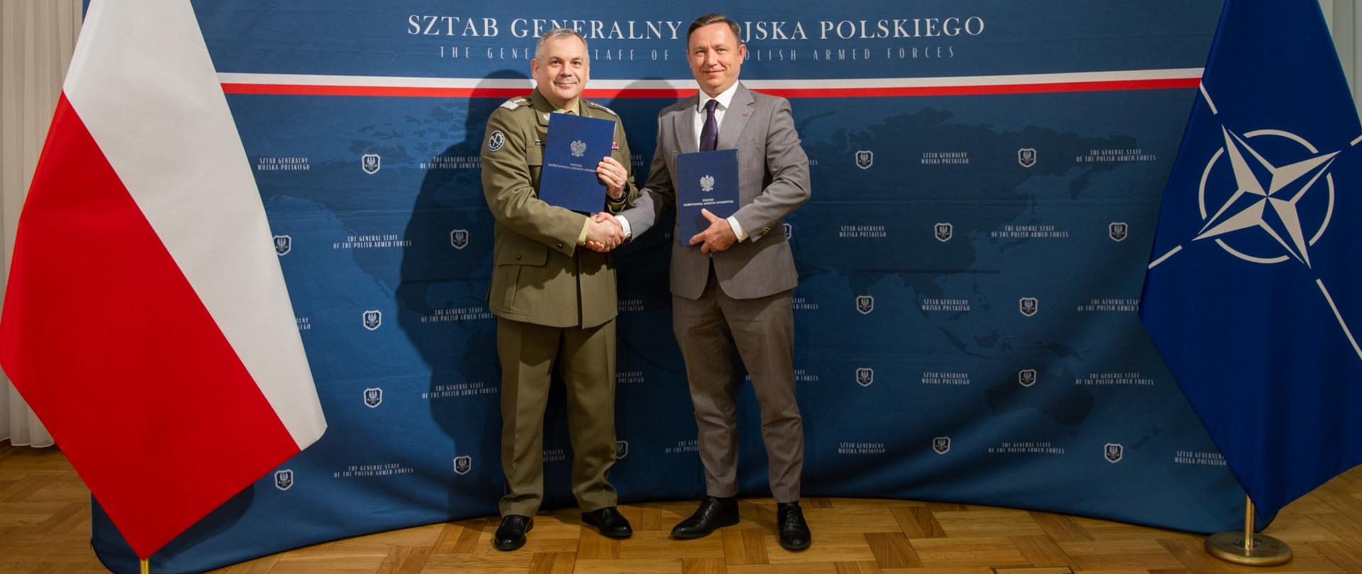 Szef Sztabu Generalnego Wojska Polskiego oraz Prezes Państwowej Agencji Atomistyki wymieniają uścisk dłoni. W ręku trzymają też po egzemplarzu podpisanego porozumienia