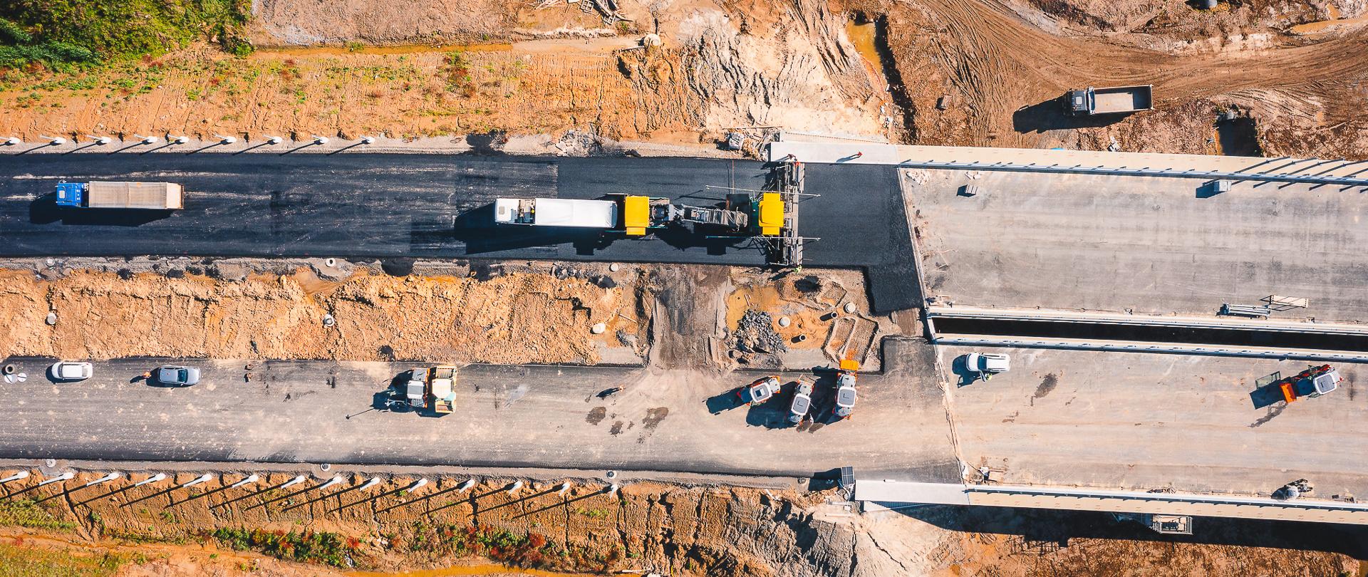 Zdjęcie przedstawia drogę ekspresową w budowie. Na zdjęciu widać plac budowy z lotu ptaka, gdzie trwa układanie asfaltu na nowej drodze. Widoczne są maszyny budowlane, ciężarówki oraz pracownicy zajmujący się pracami drogowymi. Droga jest częściowo pokryta asfaltem, a część jest jeszcze w stanie surowym. Wokół placu budowy znajdują się tereny zielone i ziemne nasypy.