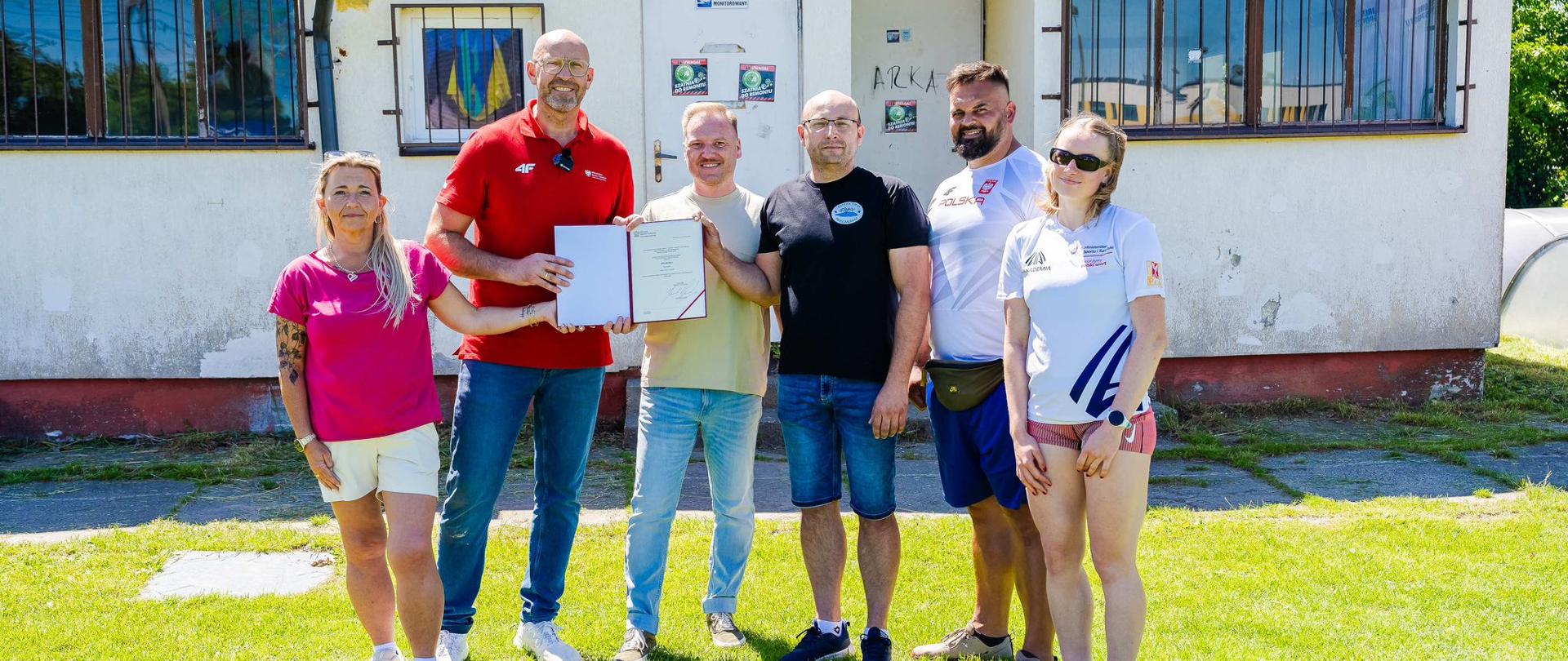Minister sportu i turystyki Jakub Rutnicki i pięć innych osób prezentują decyzję o dofinansowaniu z z programu Szatnia na medal, stojąc przed budynkiem szatni, który zostanie wyremontowany.