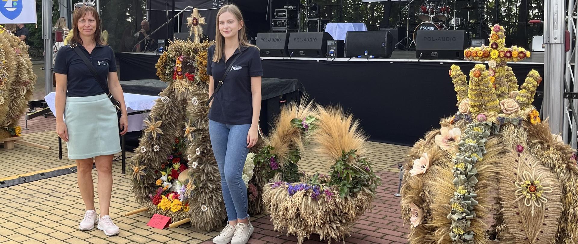 Dwie inspektorki WIORiN stoją obok tradycyjnych wieńców dożynkowych wykonanych z kłosów zbóż, kwiatów i traw. W tle znajduje się scena z zawieszonym na niej banerem, na którym widniej napis ,,Dożynki Powiatowe”, Biała Piska 13.09.2025