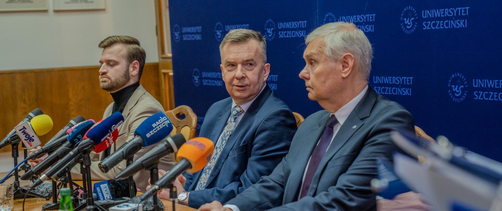 Minister Wieczorek i dwóch mężczyzn w garniturach siedzą za drewnianym stołem, za nimi niebieska ścianka z napisem Uniwersytet Szczeciński.
