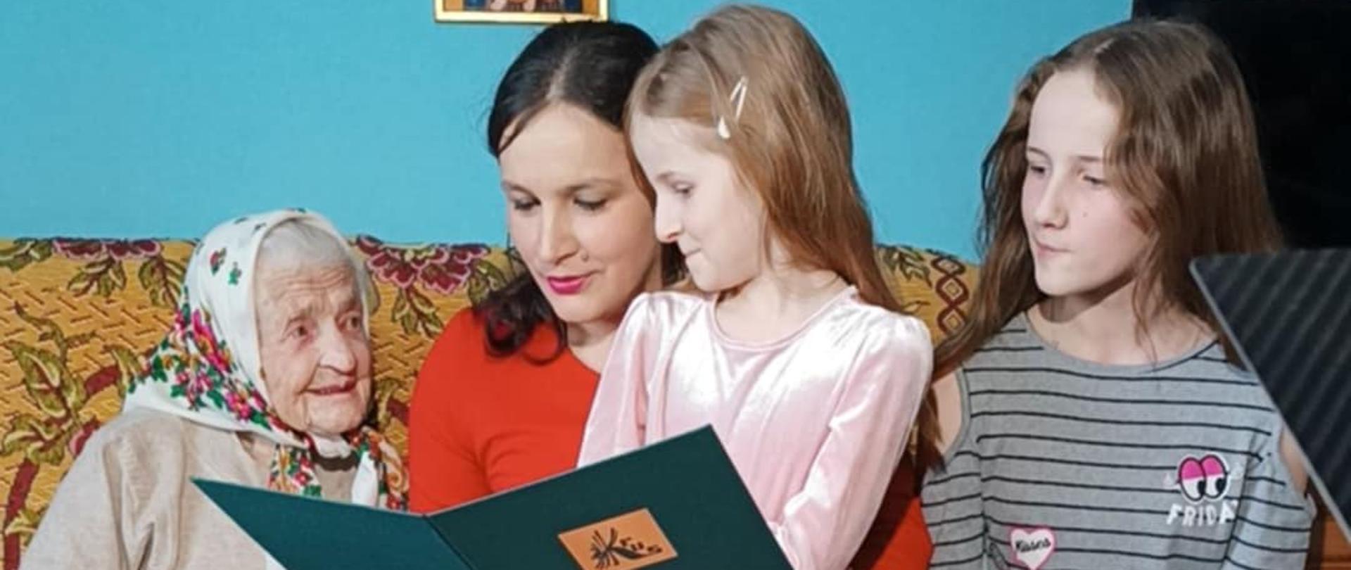 W pokoju na kanapie siedzą cztery osoby. Od lewej: Pani Anna Winiarska, jej prawnuczka (czyta list gratulacyjny od Prezesa KRUS) oraz dwie praprawnuczki.