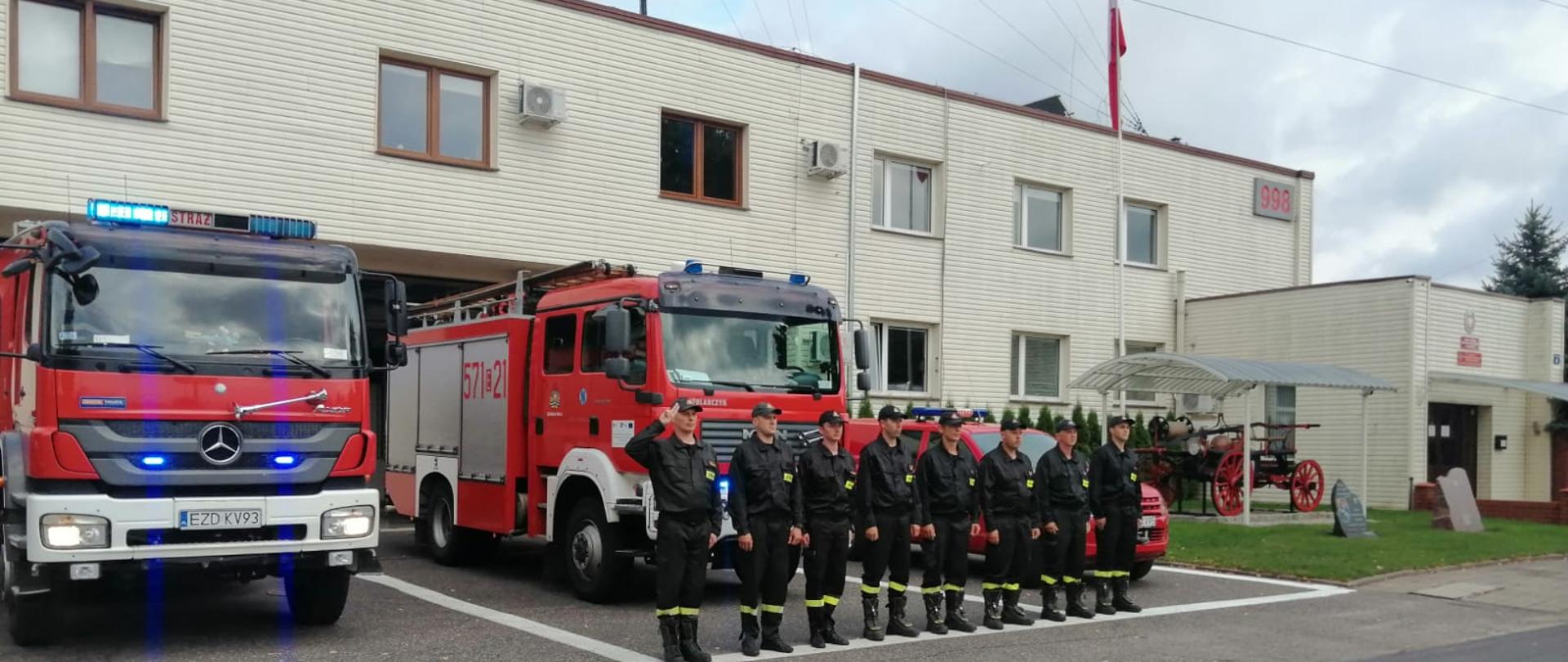 Na zdjęciu widoczni są funkcjonariusze Komendy Powiatowej PSP w Zduńskiej Woli w umundurowaniu koszarowym oraz pojazdy pożarnicze z włączoną sygnalizacją błyskową przed budynkiem garażu jednostki ratowniczo-gaśniczej . Dowódca zmiany oddaje honor bohaterom biorącym udział w walce o Niepodległą Polskę.