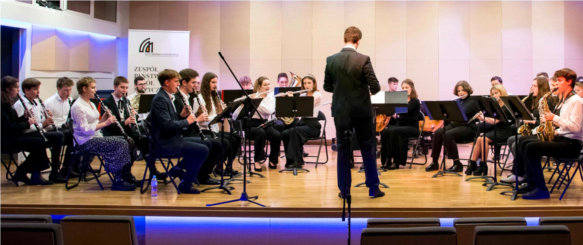 Grafika przedstawia muzykujących uczniów, tworzących orkiestrę dętą ZPSM