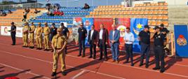 Komendant Miejski przemawia do mikrofonu za nim w rzędzie stoją zaproszone osoby, z tyłu trybuny stadionu