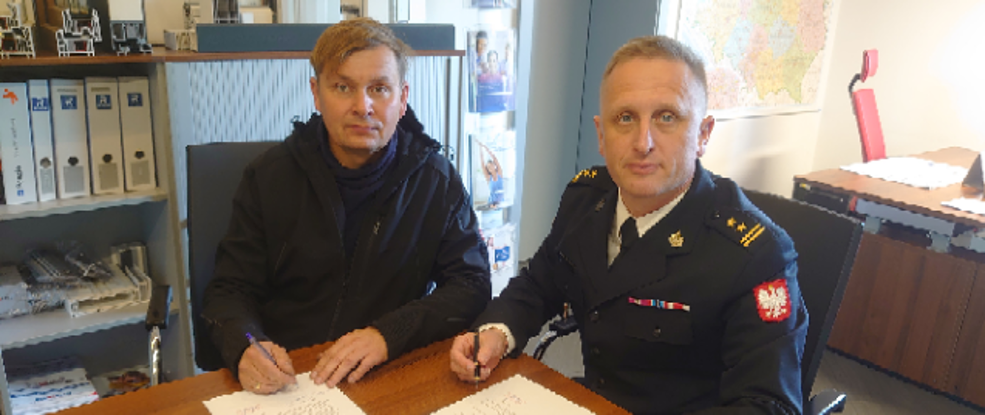 Na zdjęciu znajduje się komendant PSP w mundurze oraz prezes firmy, obie osoby siedzą przy stoliku i podpisują dokumenty