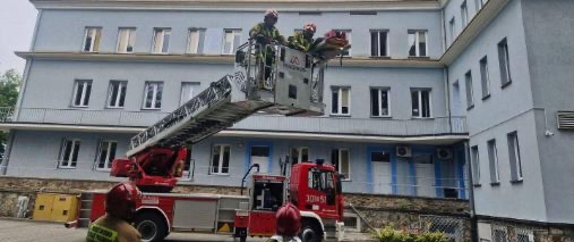 Fotografia przedstawia drabinę mechaniczną rozstawioną na placu manewrowym przy szpitalu. Na pierwszym planie 2 ratowników obserwuje pracę maszyny. W koszu urządzenia znajduje się rota zaopatrzona w nosze i przygotowana do ewakuacji poszkodowanych.