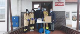 Druhny i druhowie z ochotniczej straży pożarnej w Korytowie stoją przed strażnicą. Przed nimi stoi stolik, na którym ustawione są pudełko z ulotkami i maseczkami oraz płyny do dezynfekcji