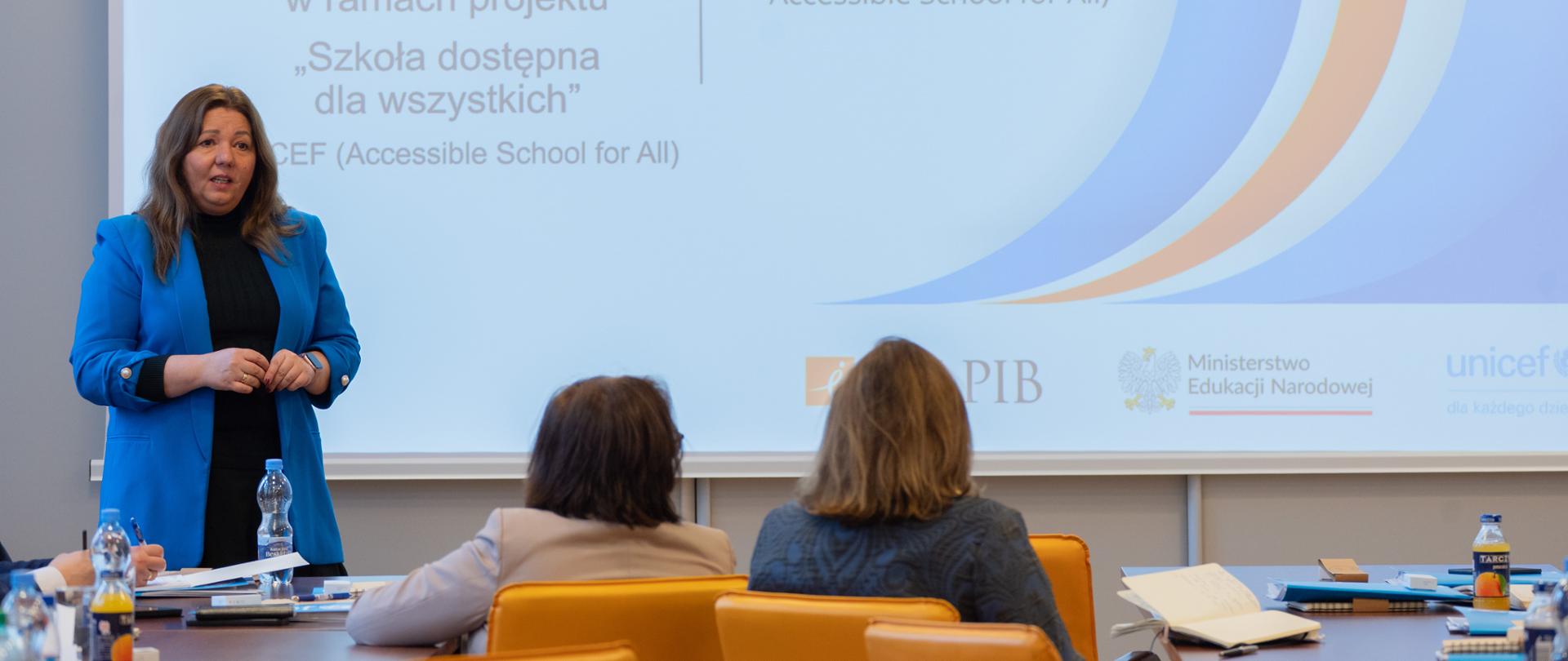 Wiceminister Izabela Ziętka stoi i przemawia do uczestników spotkania. W tle wyświetlona jest prezentacja ze szczegółami wydarzenia.
