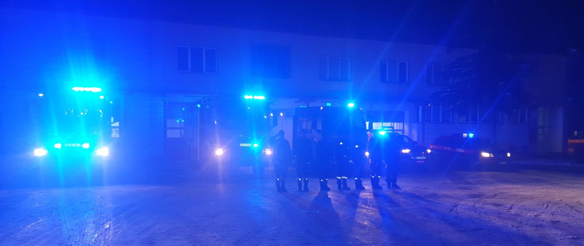 Zdjęcie wykonane w porze nocnej przedstawia grupę strażaków w czarnych ubraniach koszarowych ustawionych w szeregu przed pięcioma samochodami ratowniczo-gaśniczymi wysyłającymi niebieskie światła ostrzegawcze. Samochody ustawione na tle budynku komendy straży pożarnej w Chojnicach.