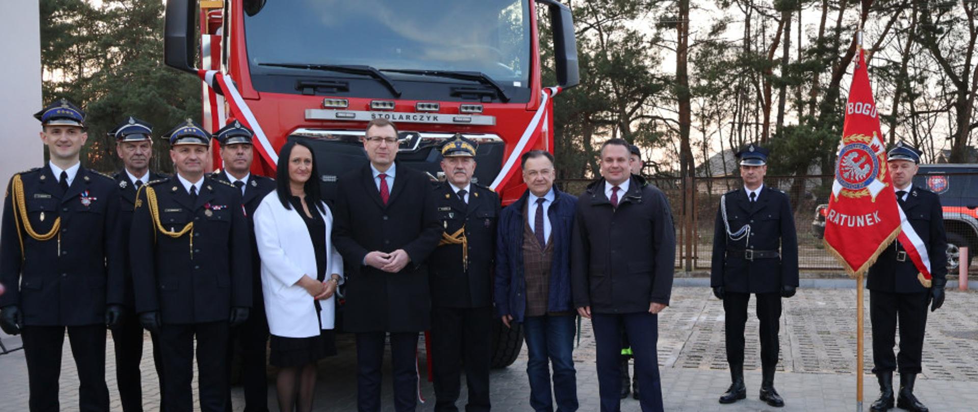 Zdjęcie grupowe gości stojących na tle samochodu strażackiego. Obok stoi strażak trzymający sztandar jednostki ochotniczej straży pożarnej. 