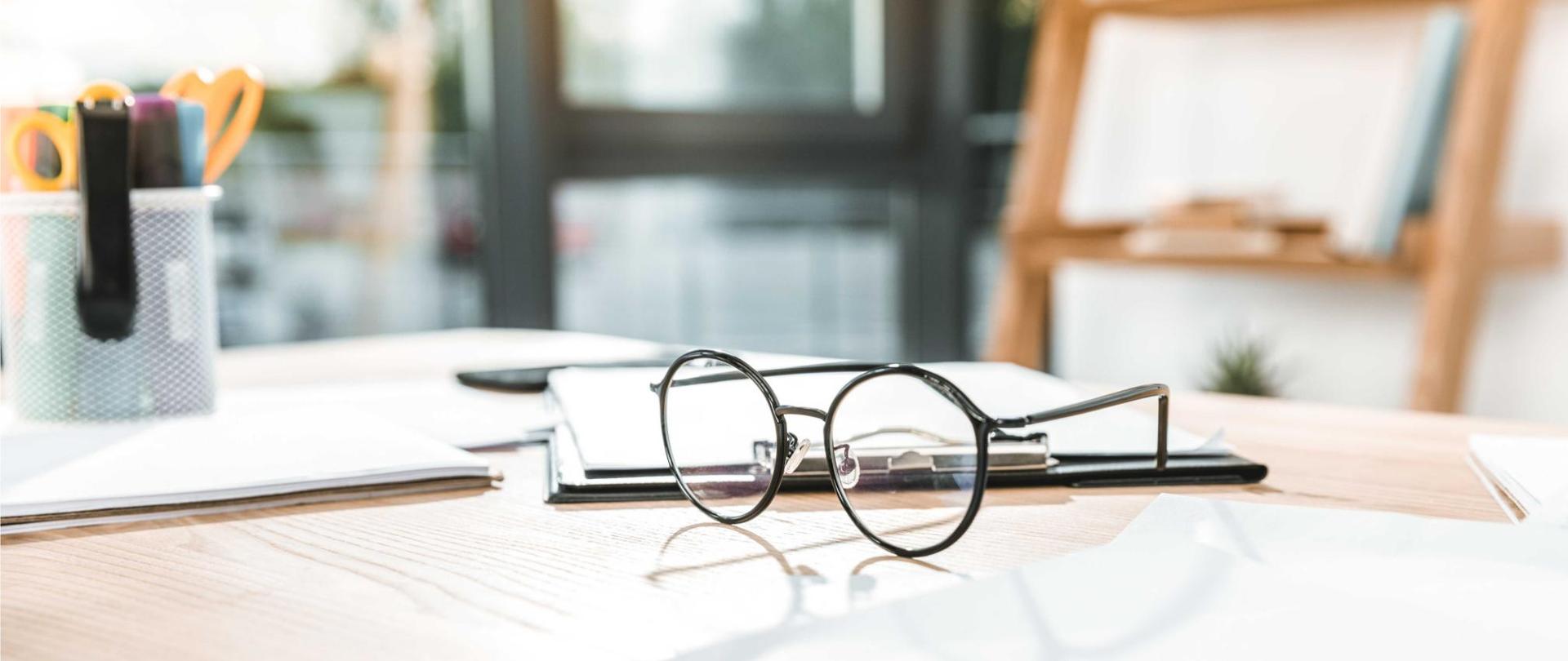 Na zdjęciu w centrum widać okulary korekcyjne leżące na biurku. W tle znajdują się przybory biurowe.