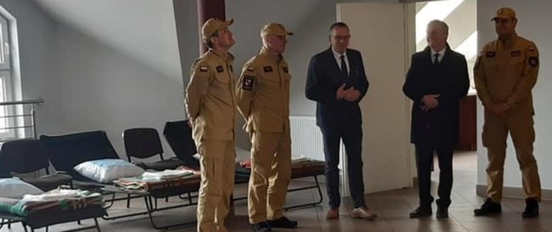 Na zdjęciu przedstawiono przygotowaną salę sypialną w remizie Ochotniczej Straży Pożarnej w Leszczynach oraz wizytujących gości. Od lewej bryg. Mariusz Góra, st. bryg. Grzegorz Rajca, zastępca wójta gminy Masłów Arkadiusz Kubiec, wojewoda świętokrzyski Zbigniew Koniusz i st. bryg. Krzysztof Ciosek. 